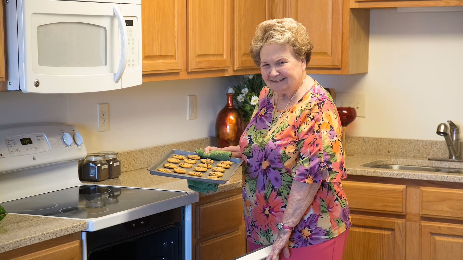 An elderly woman in a colorful floral blouse is standing in a kitchen holding a baking tray with freshly baked cookies. The kitchen has wooden cabinets, a white microwave, a stove, and a sink with a faucet.