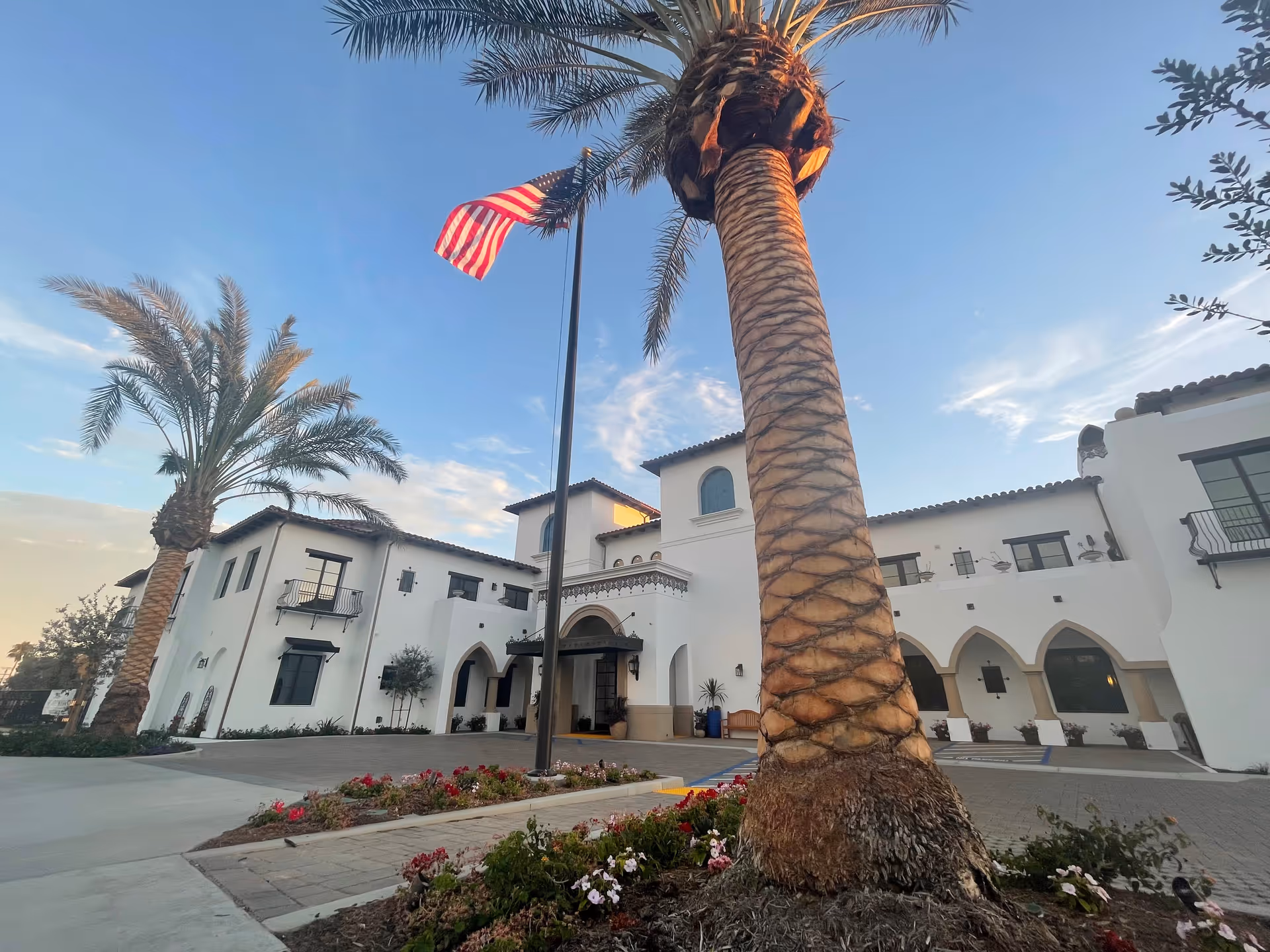 Exterior view of Raya's Paradise San Clemente facility with white stucco walls, arched doorways, and Spanish-style roof tiles. Two tall palm trees and an American flag on a flagpole are visible in the foreground, along with landscaped flower beds.