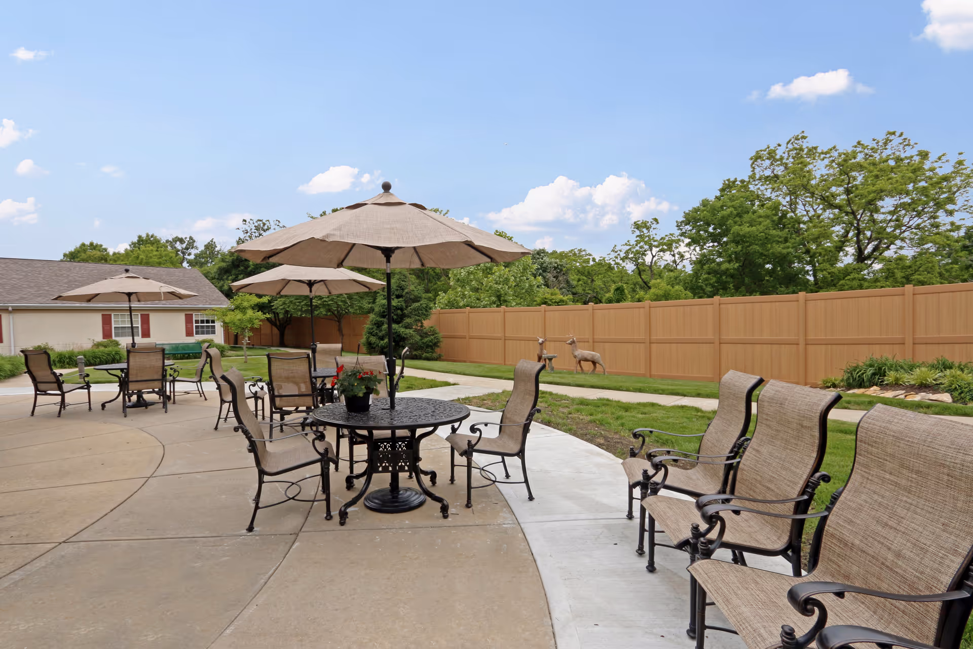 Outdoor patio area with several tables and chairs under beige umbrellas, surrounded by a concrete walkway and green grass. There is a tan privacy fence and trees in the background under a blue sky with some clouds.