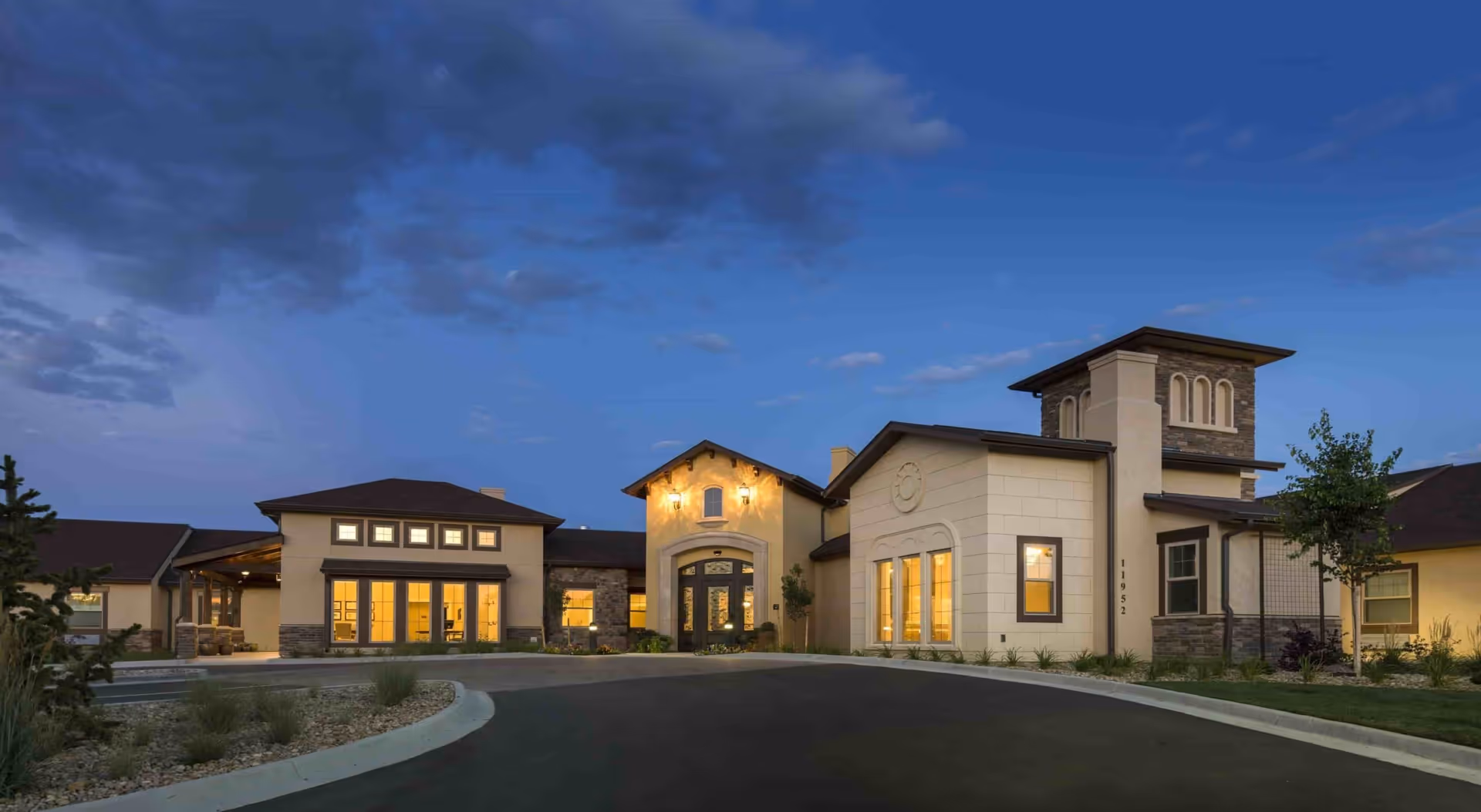 Front exterior of a senior living facility at dusk with warm interior lights and a curved driveway.