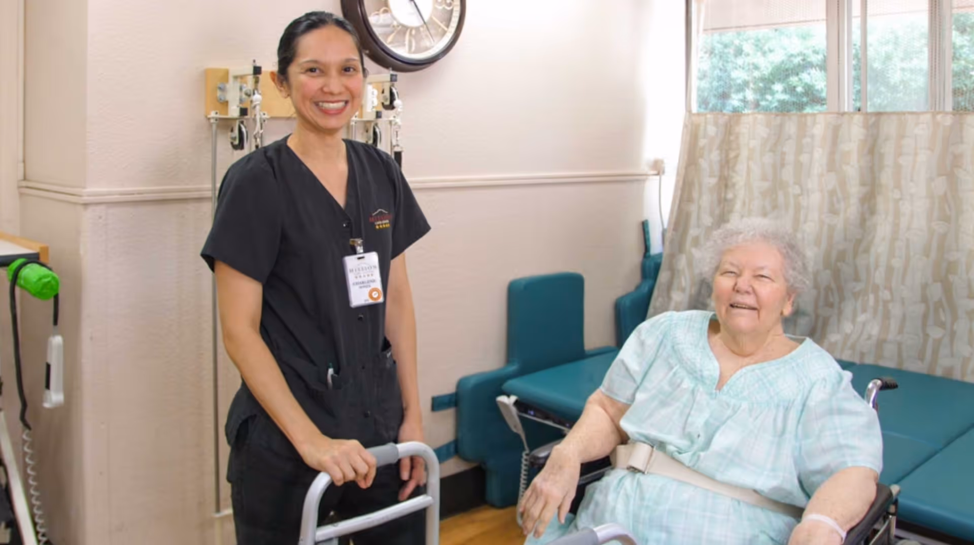 A smiling female healthcare worker in black scrubs stands next to an elderly woman seated in a wheelchair in a medical room with examination tables and medical equipment.