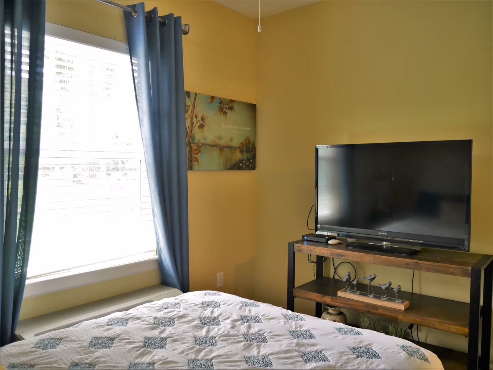 A bedroom with a bed covered in a white quilt with blue patterns, a window with blue curtains letting in natural light, a wall painting with a nature theme, and a flat-screen TV on a wooden and metal stand with decorative items underneath.