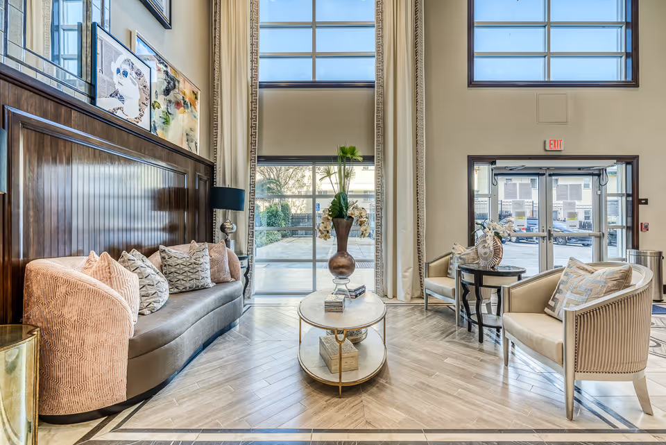 Bright and spacious senior living facility lobby with tall windows, a curved sofa with multiple cushions, two armchairs with patterned pillows, a round coffee table with a decorative vase and books, and framed artwork on the wall above the sofa.
