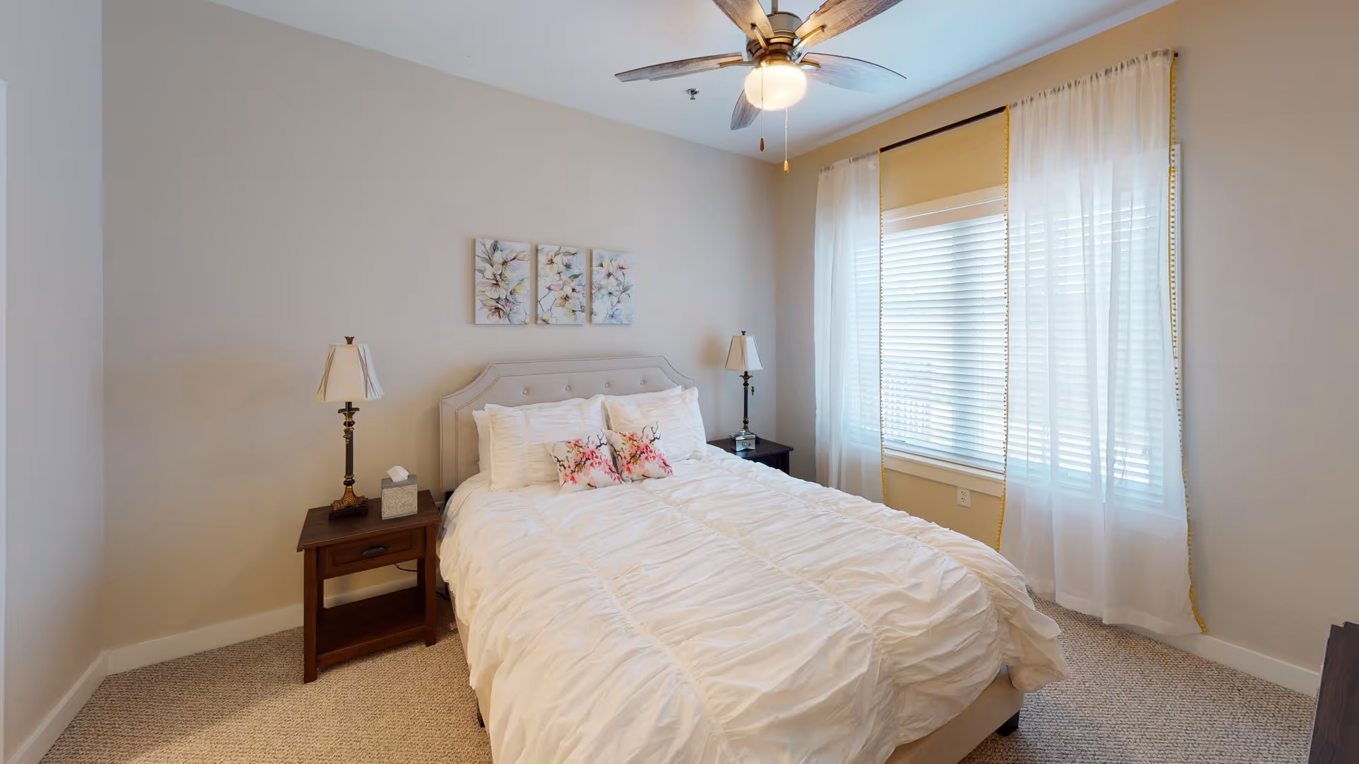 A cozy bedroom with a neatly made bed featuring white bedding and floral accent pillows. The room has beige walls, a ceiling fan with a light, two wooden nightstands each with a lamp, and a window with white sheer curtains allowing natural light to enter.