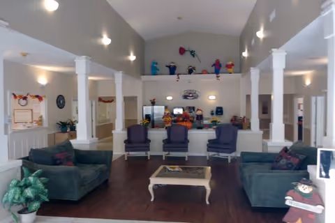 A spacious and well-lit common area in a senior living facility with two green sofas, three purple armchairs, a coffee table, and decorative plants. The room has high ceilings with wall-mounted lights and white pillars. There are colorful decorations on a ledge above a counter area in the background.