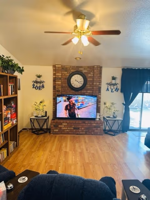 Cozy living room featuring a brick wall with a clock, a television displaying a scene, bookshelves filled with books, and decorative plants.
