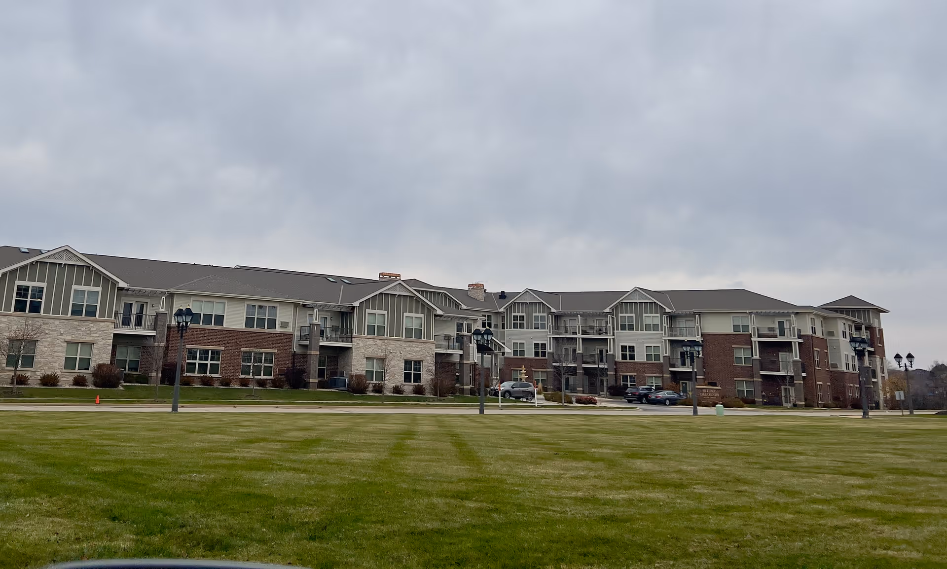 Wide exterior view of a large multi-story senior living facility building with a mix of brick and siding, multiple windows, balconies, and lamp posts on a cloudy day with a large green lawn in the foreground.