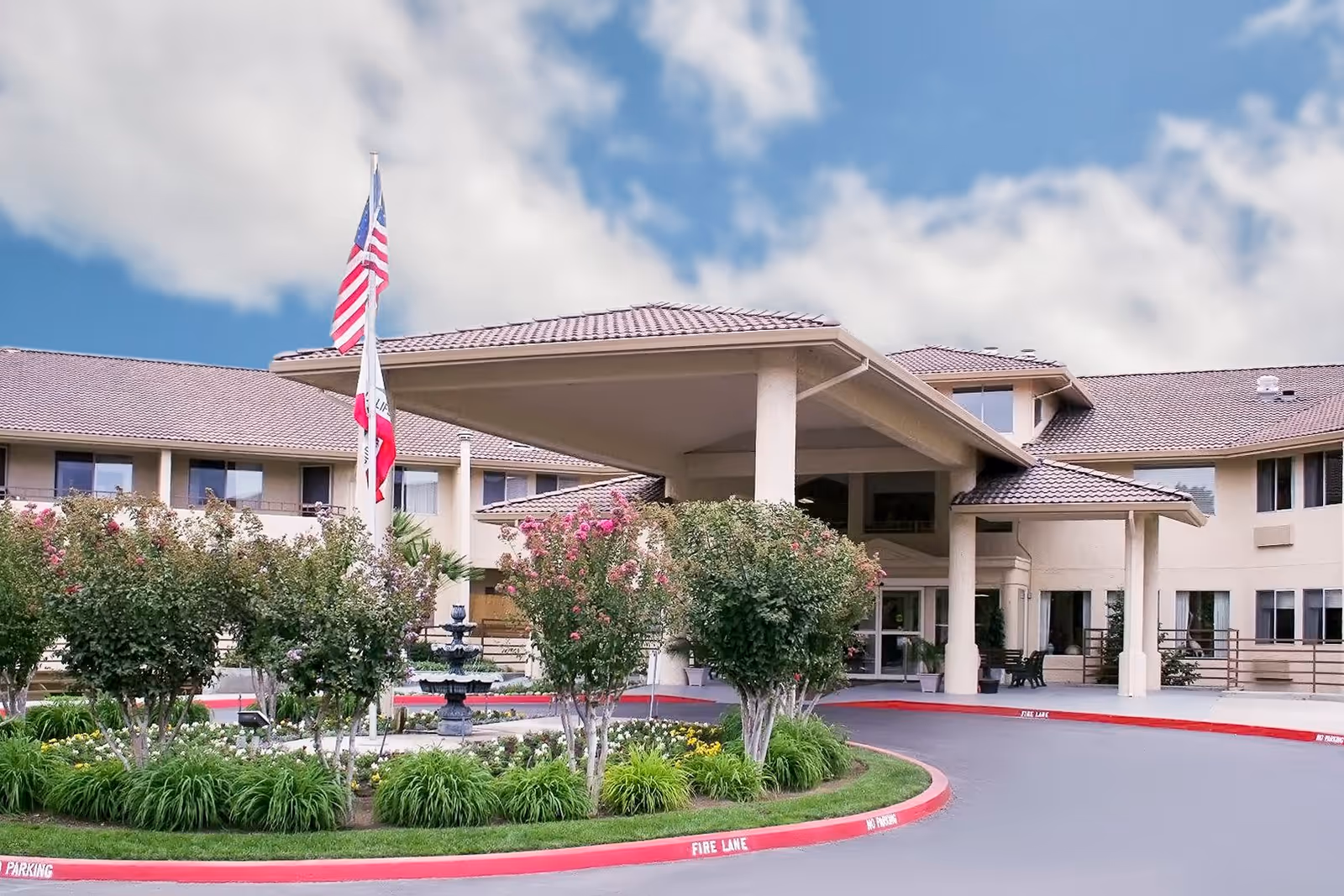 Exterior view of Truewood by Merrill, Fig Garden senior living facility showing the main entrance with a covered driveway, landscaped garden with flowering bushes and a fountain, and American and California state flags on flagpoles against a partly cloudy sky.