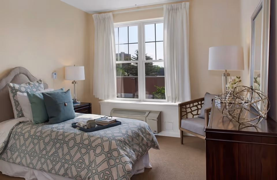 Well-lit furnished bedroom with a bed, bedside tables, a window, and a dresser with decorative items.