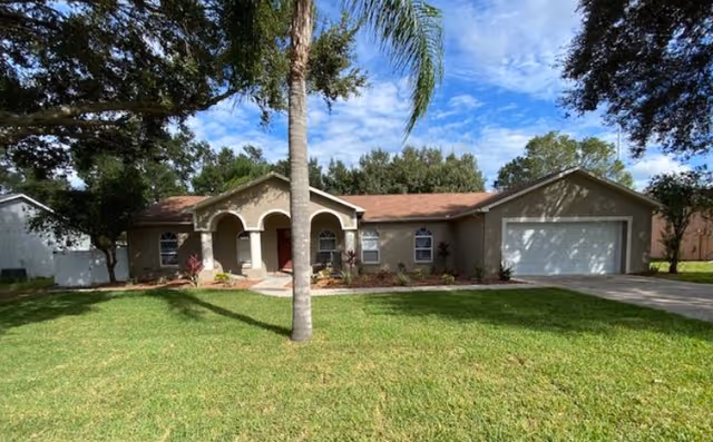 Single-story house with a beige exterior, a red front door, and a two-car garage. The house is surrounded by a well-maintained green lawn with a palm tree in the foreground and other trees around the property under a partly cloudy blue sky.