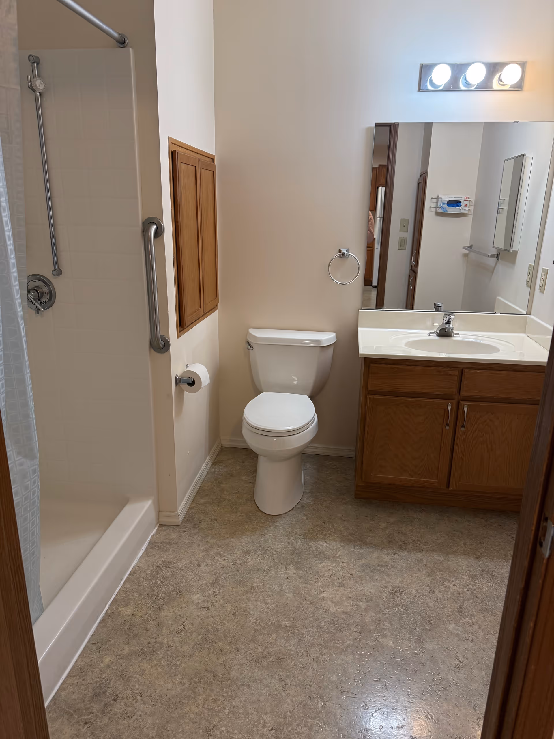 A clean bathroom with a white toilet, a wooden vanity with a sink and mirror above it, and a shower with a grab bar and shower curtain. The floor is covered with a light-colored linoleum, and the walls are painted white.