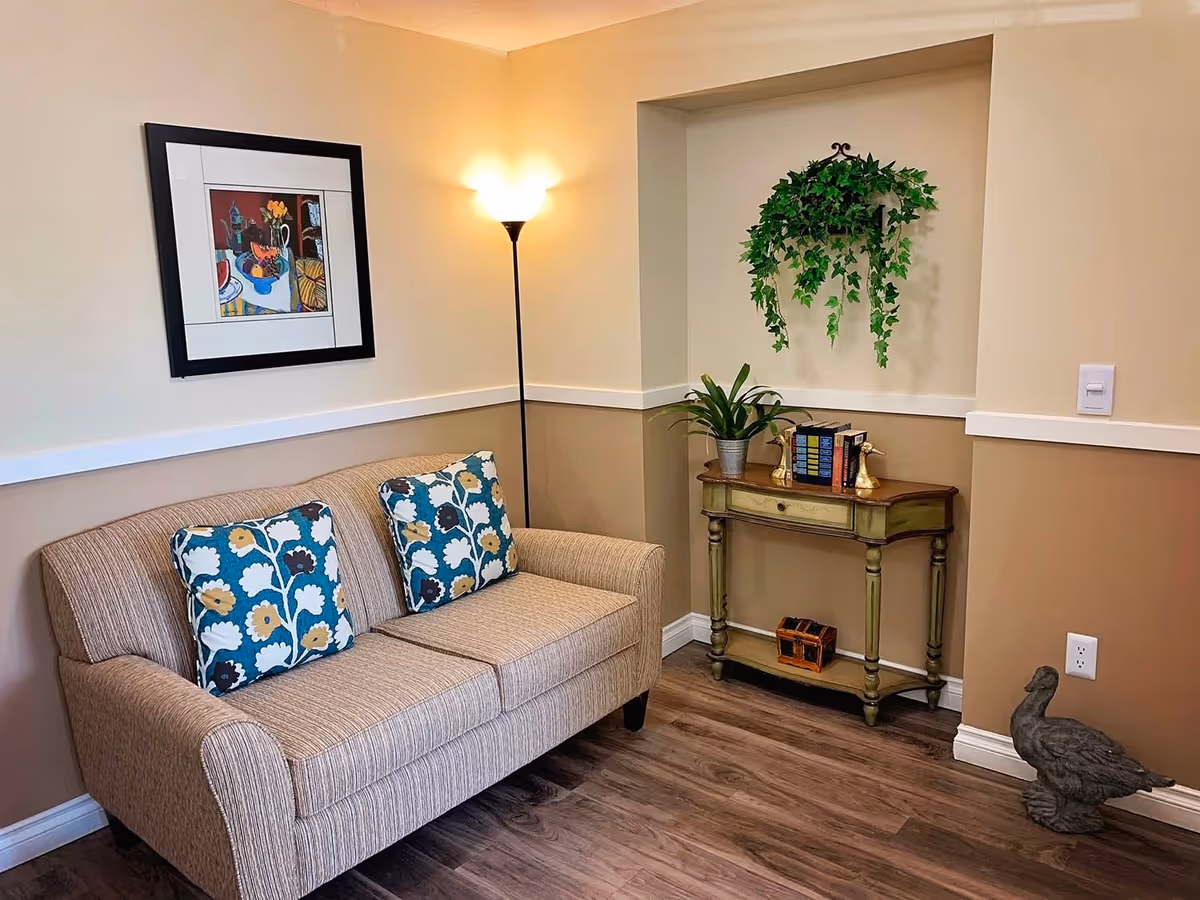 A cozy corner of a senior living facility featuring a beige upholstered sofa with two floral patterned cushions, a tall floor lamp emitting warm light, a small green wooden console table with decorative plants and books, a hanging green ivy plant in a recessed wall niche, and a duck-shaped sculpture on the wooden floor.