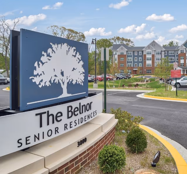 Entrance sign for The Belnor Senior Residences with a large tree logo, situated near a curved driveway and parking area. In the background, a multi-story residential building with brick and siding exterior is visible under a partly cloudy sky.
