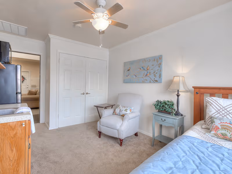 A cozy bedroom with a wooden bed partially visible on the right, covered with a blue blanket and decorative pillows. Next to the bed is a small blue nightstand with a lamp and a plant. A comfortable beige armchair with a floral pillow sits against the wall, accompanied by a small wooden side table. Above the chair is a wall art piece featuring birds on branches. The room has beige carpet, white walls, a ceiling fan with a light, and a double-door closet. A doorway leads to another room with a sofa visible.