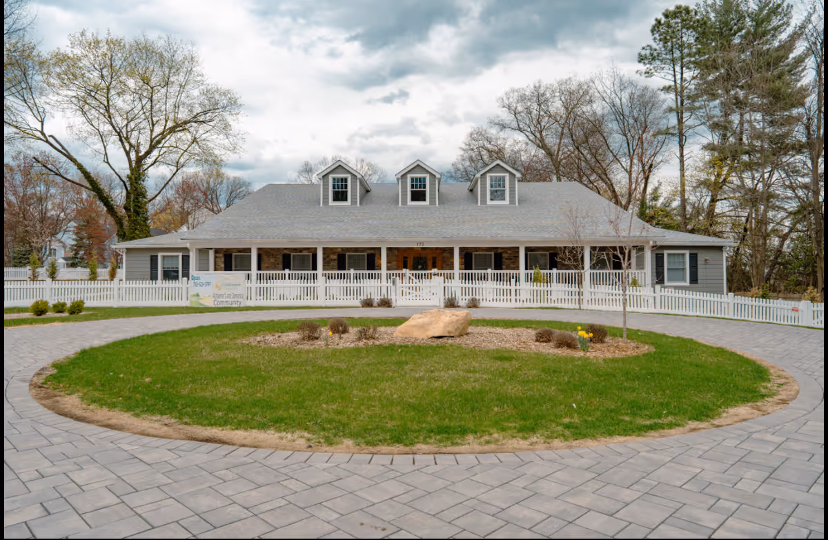 Front exterior of a single-story memory care building with a wraparound porch, white picket fence, and circular driveway around a grassy island.