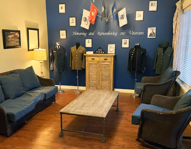 A seating room with sofas and wicker chairs around a wooden coffee table and a blue wall memorial display of military uniforms, flags, and framed photos honoring veterans.