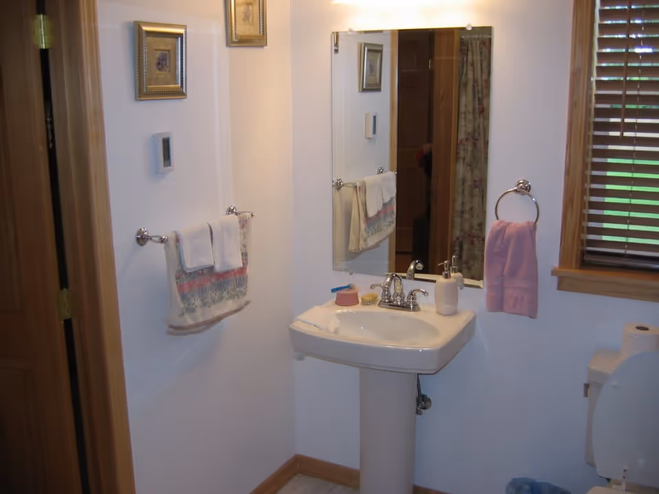 A small bathroom with a white pedestal sink, a rectangular mirror above it, and a towel rack with folded towels on the left wall. There is a pink hand towel hanging on a ring next to a window with wooden blinds. A toilet with the lid open is partially visible on the right side, along with a roll of toilet paper on the tank. The walls are white and there are two small framed pictures hanging above the towel rack.