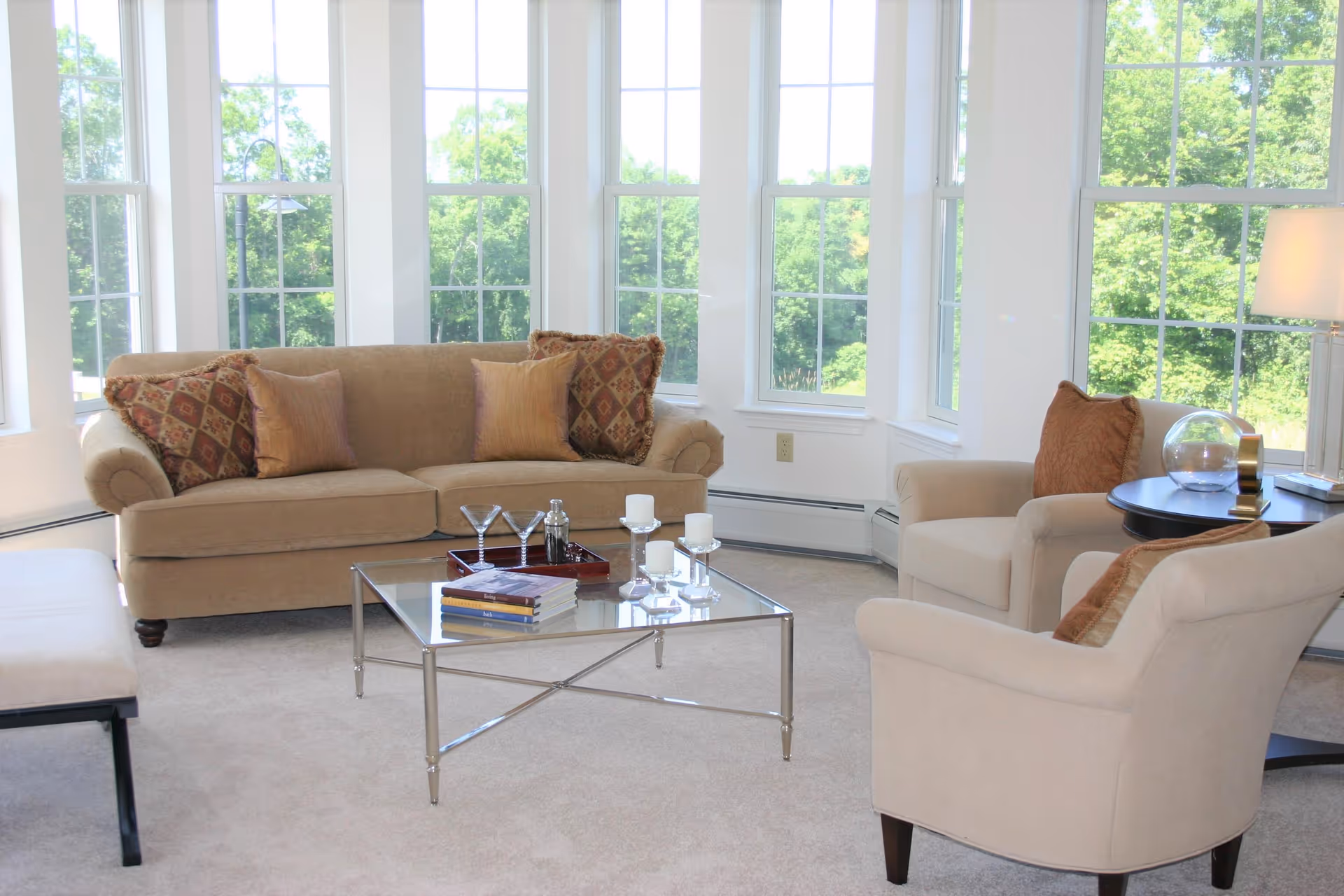 A bright living room with large windows showing green trees outside. The room features a beige sofa with patterned and solid pillows, two beige armchairs with pillows, a glass coffee table with books, candles, and a tray with martini glasses, and a side table with a lamp and decorative items.