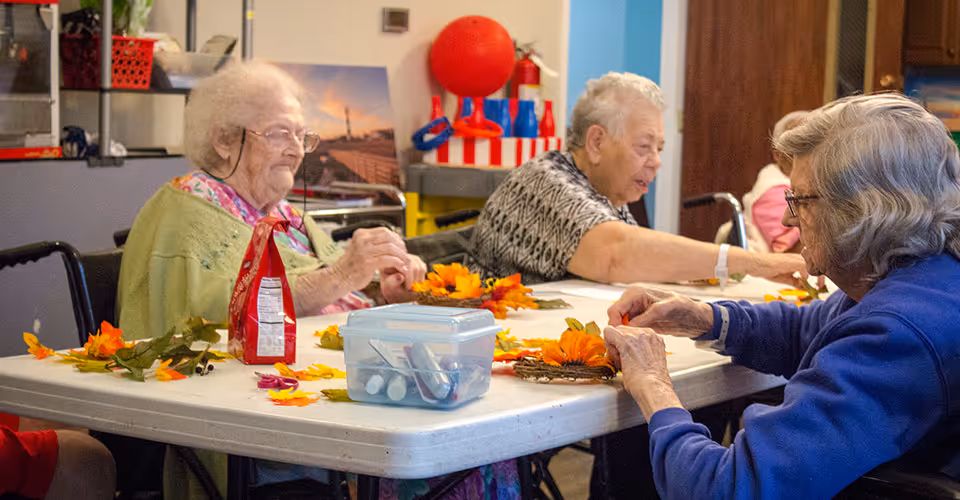 Three elderly women sitting around a table engaged in a craft activity involving artificial flowers and leaves. The table has a plastic container with glue sticks and other craft supplies. The setting appears to be a communal room with shelves and various items in the background.