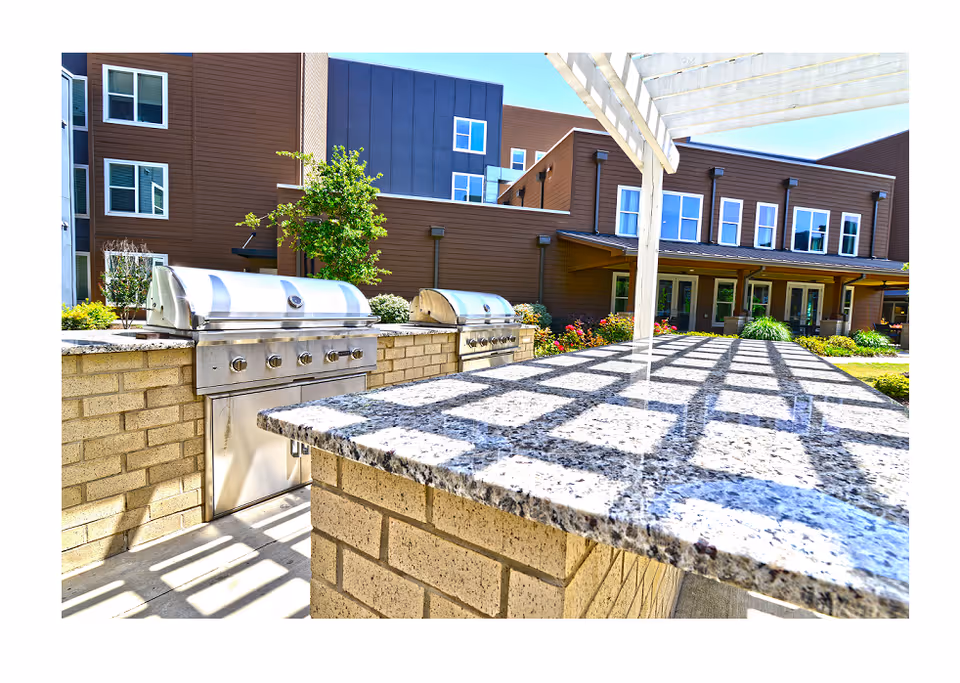 Outdoor courtyard barbecue area showing stainless steel built-in grills and a granite countertop beneath a pergola, with the facility building and landscaping in the background.