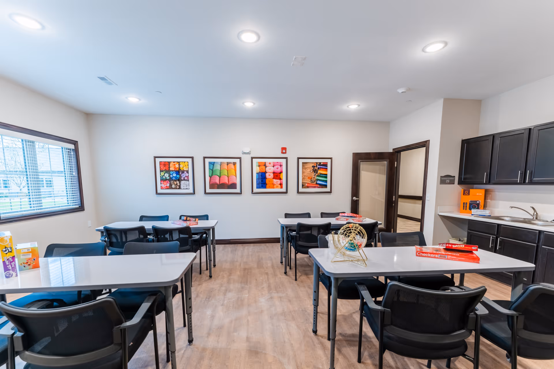 A bright activity room with several tables and black chairs arranged neatly. The room has light-colored walls with four colorful framed pictures of yarn and knitting supplies on one wall. There is a large window on the left side letting in natural light. On the right side, there is a kitchenette area with dark cabinets, a sink, and some board games like Checkers and Yahtzee placed on the tables.