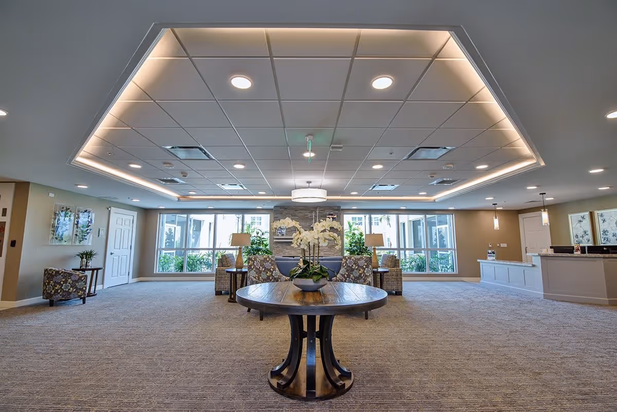 Bright, spacious lobby with a central round table holding orchids, seating areas, and a reception desk under recessed ceiling lighting.