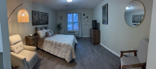 A cozy bedroom with a bed covered in a beige comforter, a nightstand with a lamp and clock, a cushioned armchair, a wooden dresser with a TV on top, a round mirror on the wall, and a window with curtains letting in natural light.
