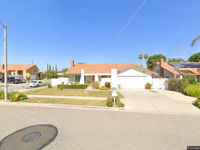 Single-story residential house with a red-tiled roof, white exterior walls, a driveway leading to a two-car garage, and a front yard with grass and shrubs under a clear blue sky.