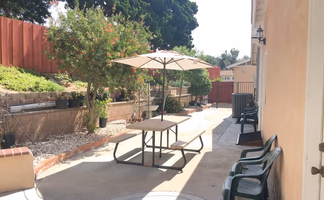 Outdoor patio area with a picnic table and umbrella, surrounded by potted plants and greenery along a retaining wall. Several plastic chairs are lined up against the building wall on the right side.