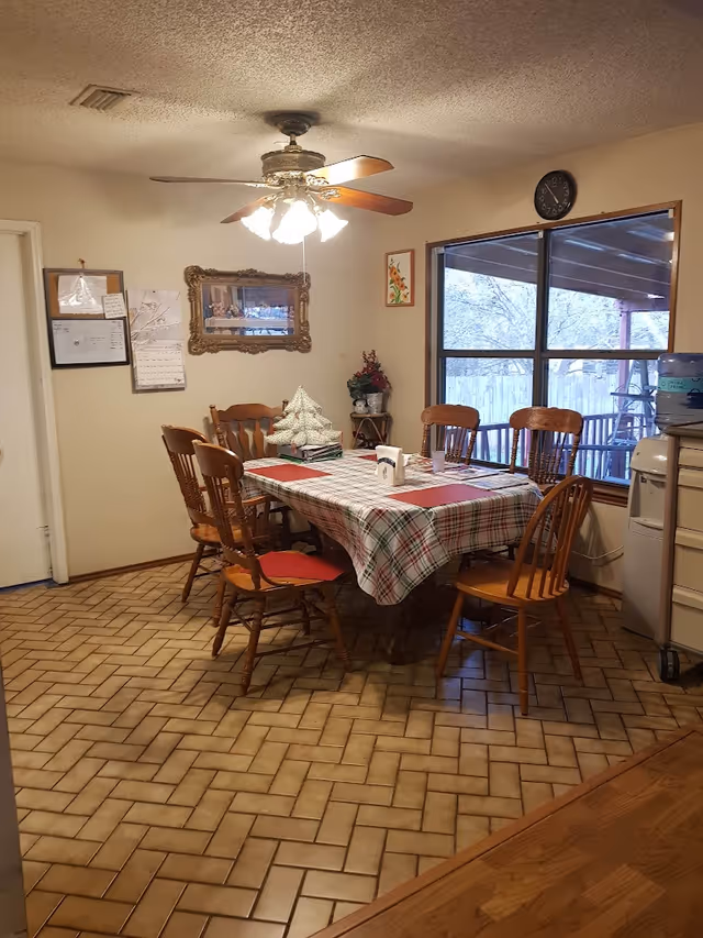 A dining room with a rectangular table covered with a plaid tablecloth and six wooden chairs around it. There is a ceiling fan with lights above the table. On the table, there are placemats, a small decorative Christmas tree, and some papers. The room has a large window showing an outdoor covered patio area. The floor is tiled in a herringbone pattern, and there are framed pictures and a clock on the walls.