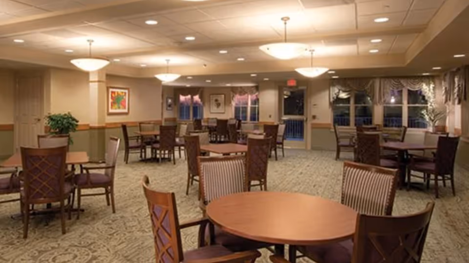 A spacious dining room with multiple round and square wooden tables surrounded by cushioned chairs. The room is warmly lit with ceiling lights and has large windows with decorative curtains. There is a plant in the corner and framed artwork on the walls.