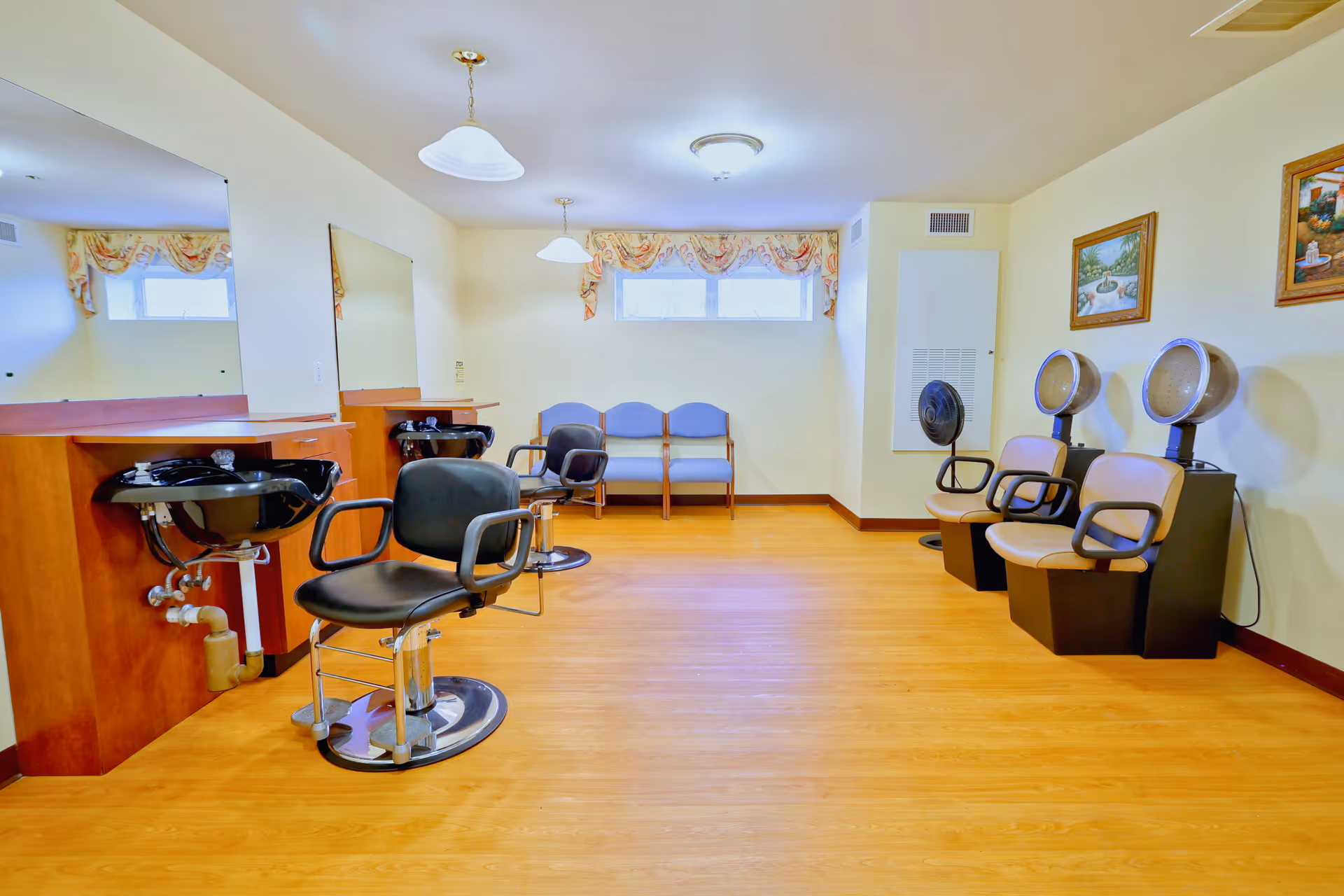 Interior of a hair salon area in an assisted living facility featuring two black salon chairs in front of sinks and mirrors on the left, two beige salon chairs with hair dryers on the right, and a row of three blue chairs against the back wall under windows with floral valances. The room has light yellow walls, wood flooring, and framed paintings on the right wall.