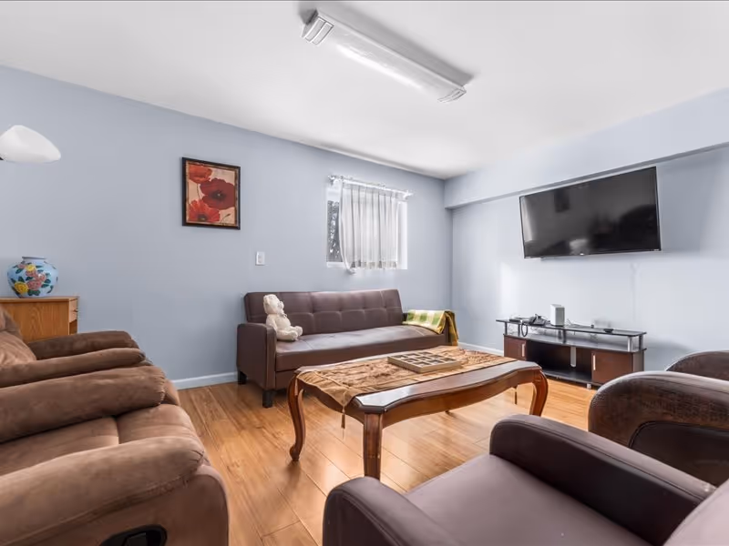 A cozy living room with light blue walls, a brown sofa with a teddy bear on it, two brown recliner chairs, a wooden coffee table with a decorative runner, a flat-screen TV mounted on the wall, a small window with sheer curtains, a framed floral painting, and a wooden cabinet with a decorative vase.