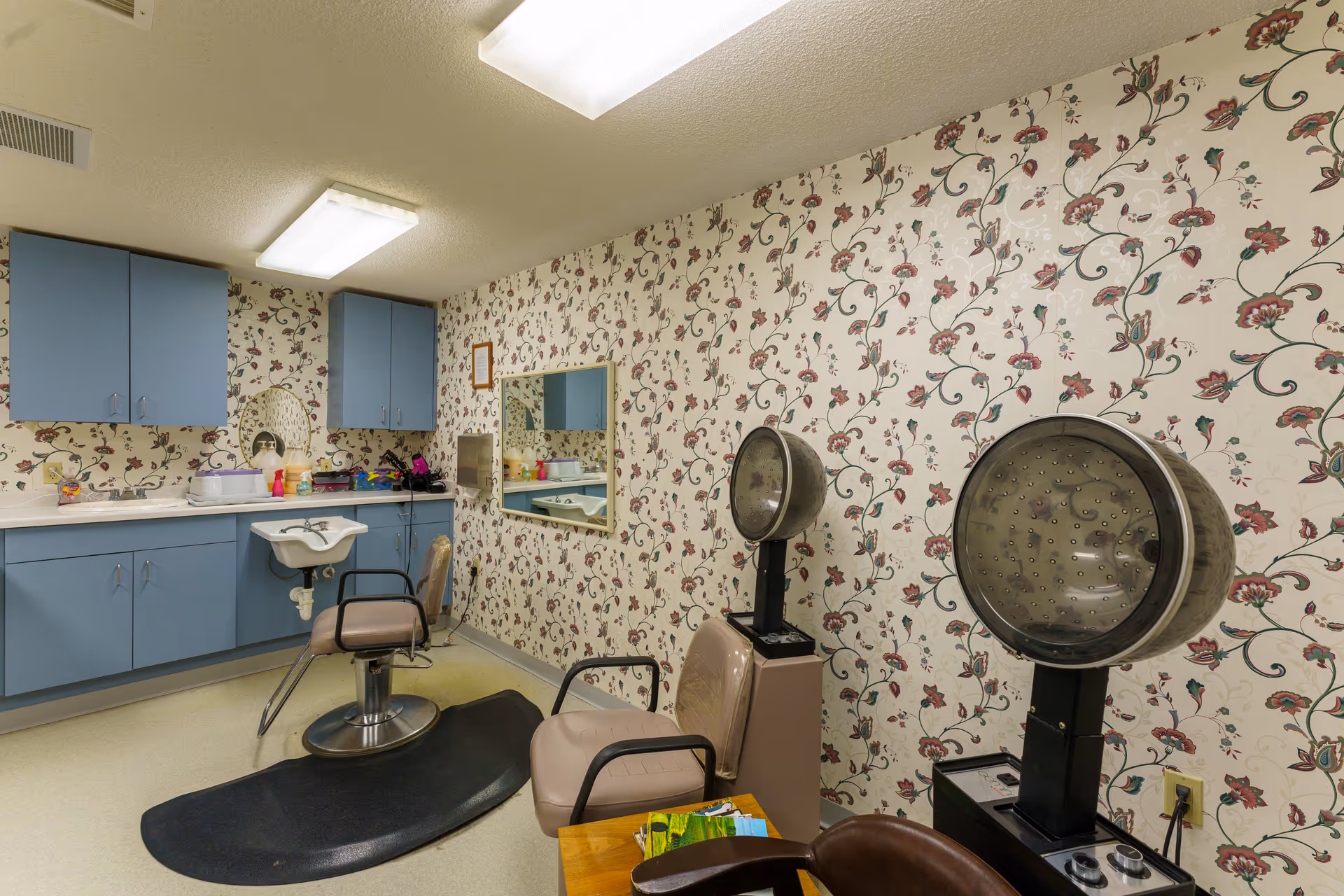 A small salon room with vintage hooded hair dryers, salon chairs, a sink and blue cabinets against floral wallpaper.