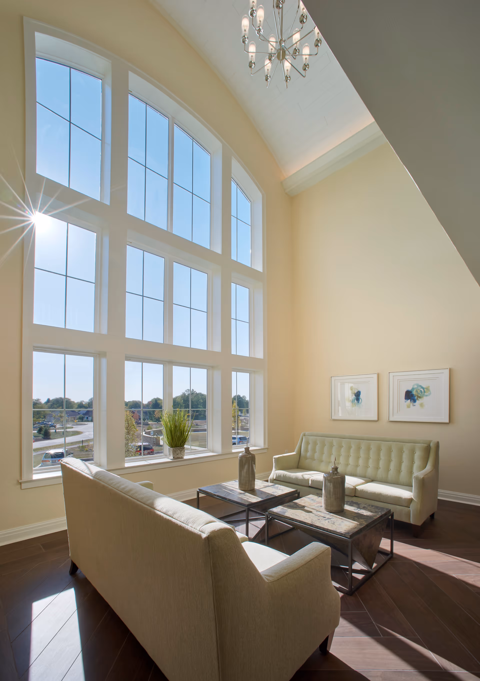 A bright living room area with large, tall windows letting in natural sunlight. The room features two beige sofas facing each other with two matching coffee tables in between. There are decorative vases on the tables and two framed abstract artworks on the wall. The floor is dark wood, and a chandelier hangs from the high ceiling.