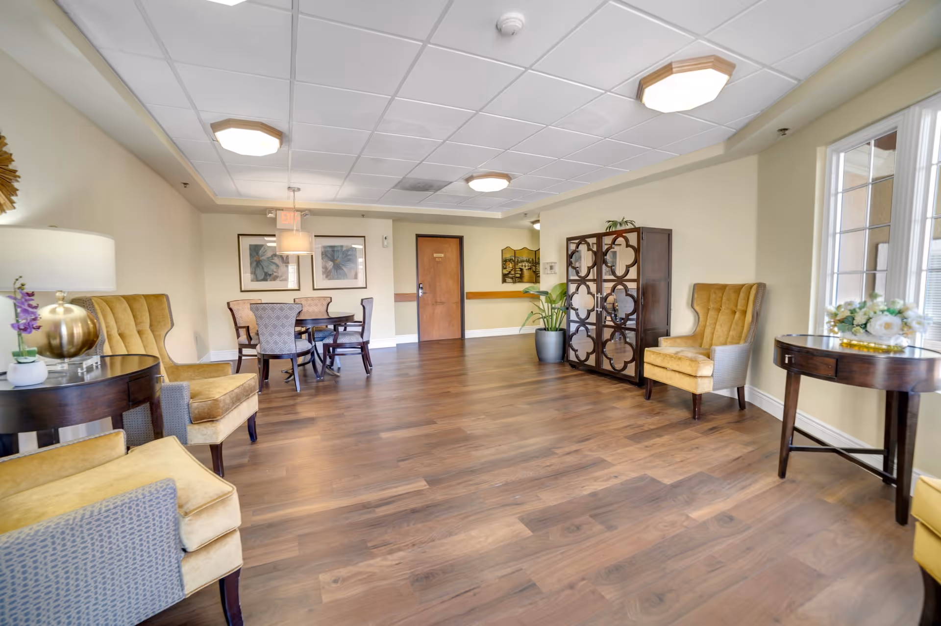 A spacious, well-lit common area with wooden flooring, beige walls, and a white ceiling with recessed lighting. The room features several upholstered armchairs in yellow and gray, a round wooden table with four chairs, decorative plants, framed artwork on the walls, and large windows letting in natural light.