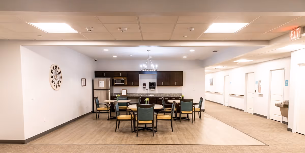 A communal dining area in a senior living facility featuring a small kitchen with dark cabinets, a refrigerator, microwave, and a granite countertop. There are two round tables with chairs arranged around them. The room has light-colored walls, a large decorative clock on one wall, and a chandelier-style light fixture above the tables. A hallway with several doors and an exit sign is visible to the right.