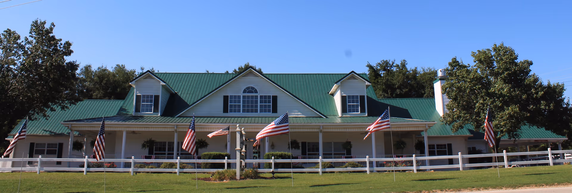 Wide house with a green metal roof, wraparound porch, white fence, and American flags on the lawn.