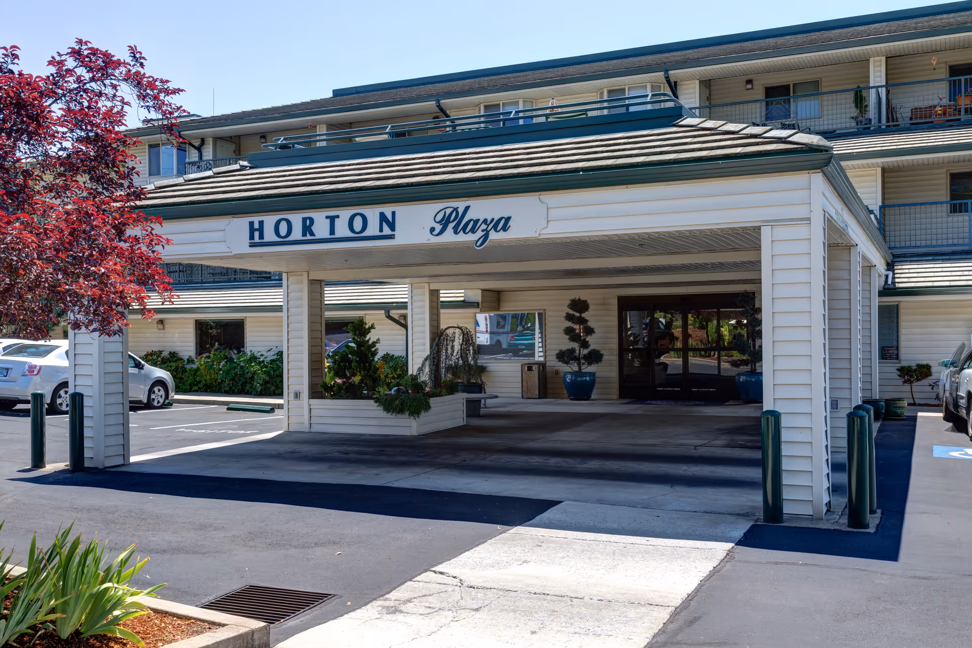 Entrance to Horton Plaza senior living facility with a covered drop-off area, potted plants, and a parking lot with cars visible on a sunny day.