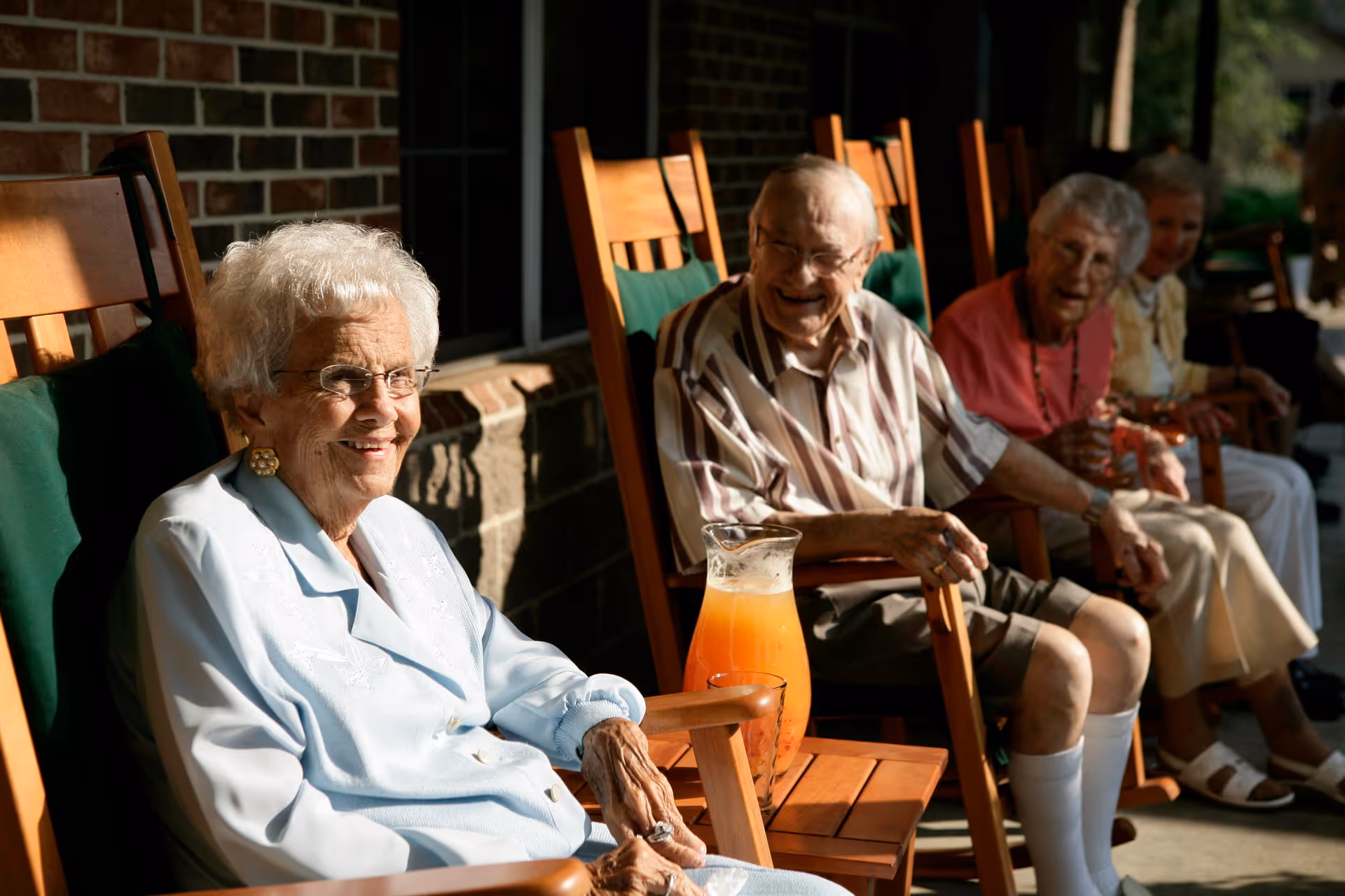 A group of elderly people sitting on wooden rocking chairs on a porch outside a brick building, enjoying a sunny day. A pitcher of orange juice and a glass are placed on a small wooden table between two of the chairs.
