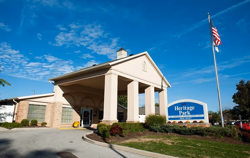 Exterior view of the Heritage Park senior living facility building with a covered entrance supported by large columns, a landscaped area with bushes and flowers, an American flag on a flagpole, and a blue sign displaying the facility name and contact number.