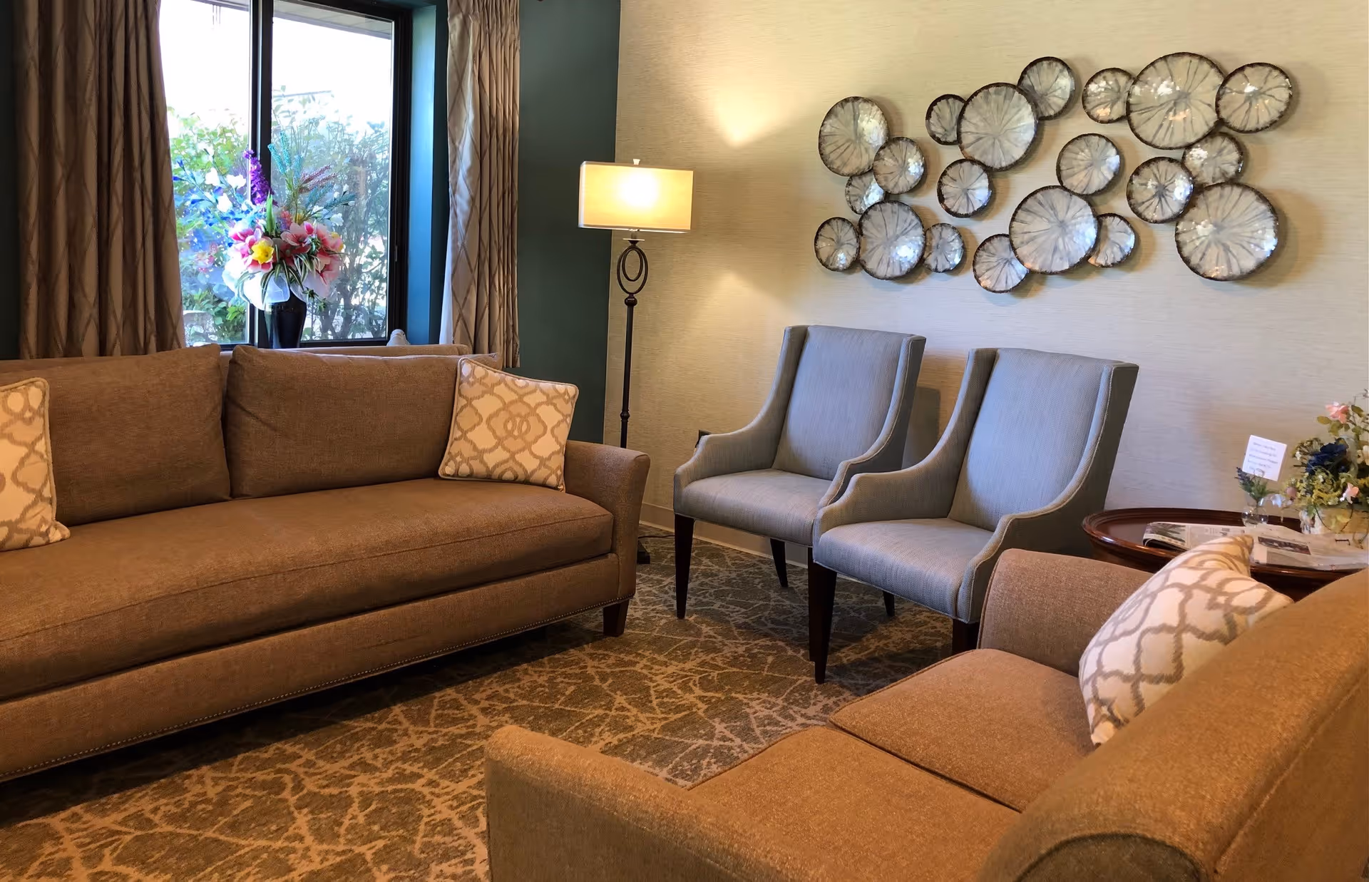 A cozy living room area with two brown sofas, two light blue armchairs, a floor lamp, a window with curtains, a decorative wall art piece made of circular metallic elements, and a side table with a flower arrangement and magazines.