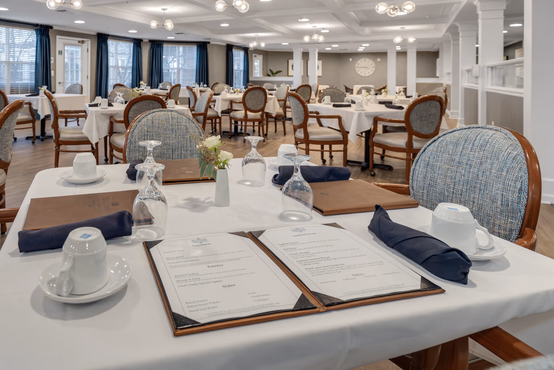 A bright and elegant dining room with multiple tables covered in white tablecloths, each set with menus, upside-down wine glasses, navy blue cloth napkins, white coffee cups on saucers, and small floral centerpieces. The room features wooden chairs with cushioned seats and backs, large windows with blue curtains, and modern ceiling light fixtures.