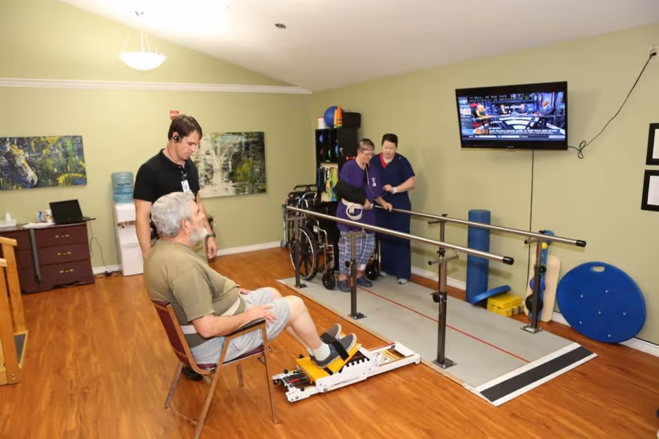 Two staff assist a resident walking with parallel bars while another resident sits using a leg exercise machine in a therapy room.