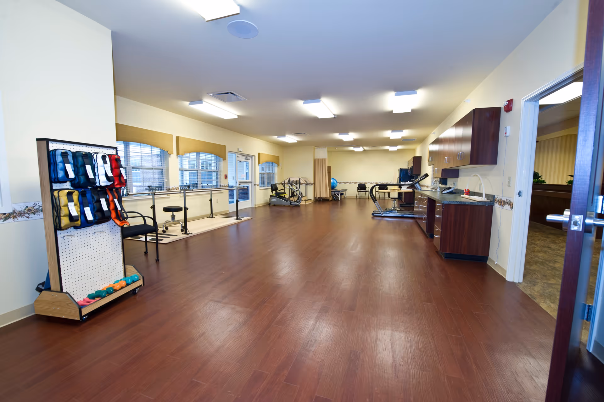 Spacious senior living rehabilitation/exercise room with hardwood floors, parallel bars, exercise equipment and cabinets along one wall.