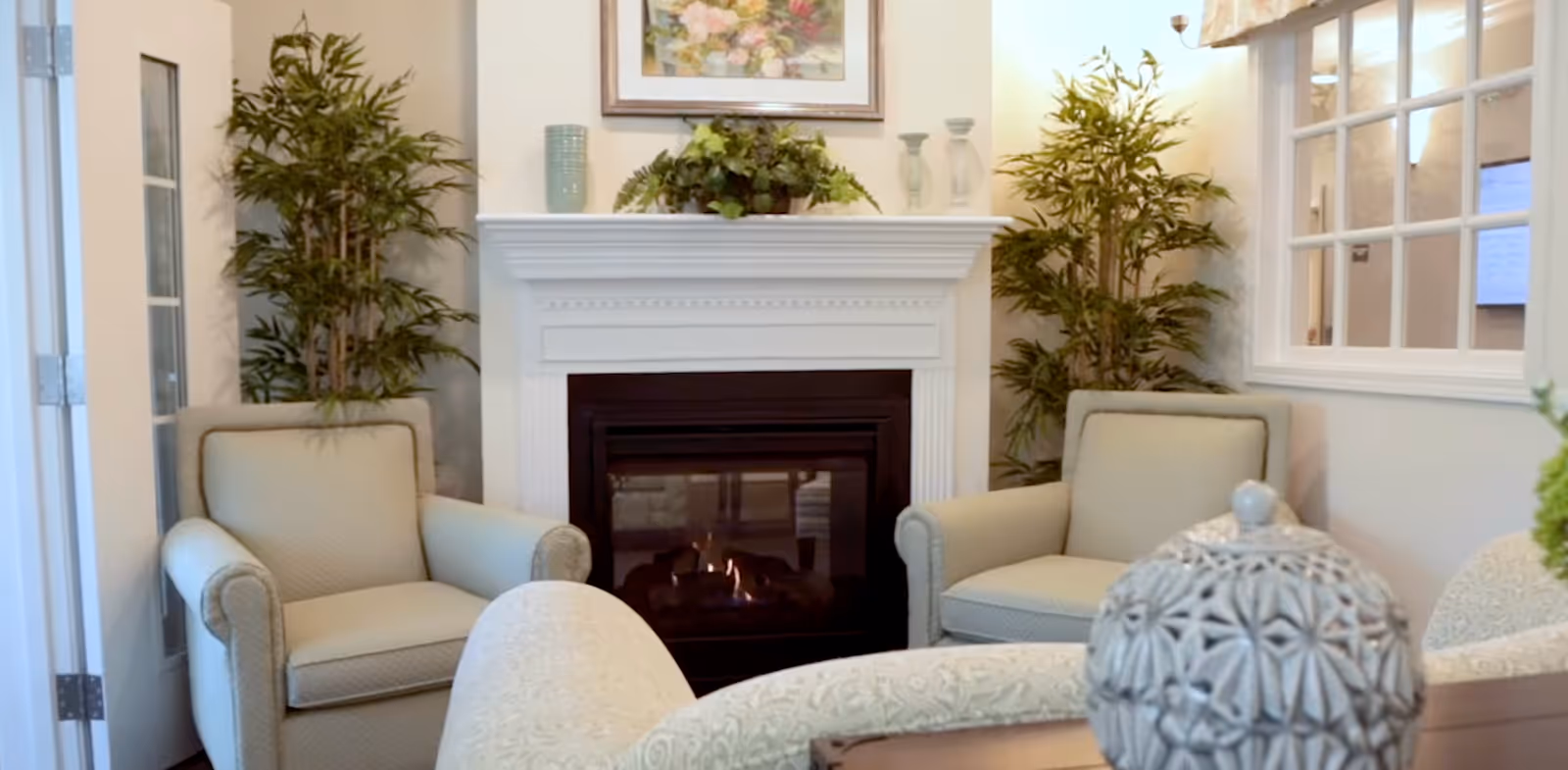 A cozy sitting area with two beige armchairs positioned on either side of a white fireplace with a fire burning inside. The fireplace mantel is decorated with a plant, two glass candle holders, and a blue vase. There are two tall green plants flanking the fireplace, a framed floral painting above it, and a window with a floral valance on the right side.
