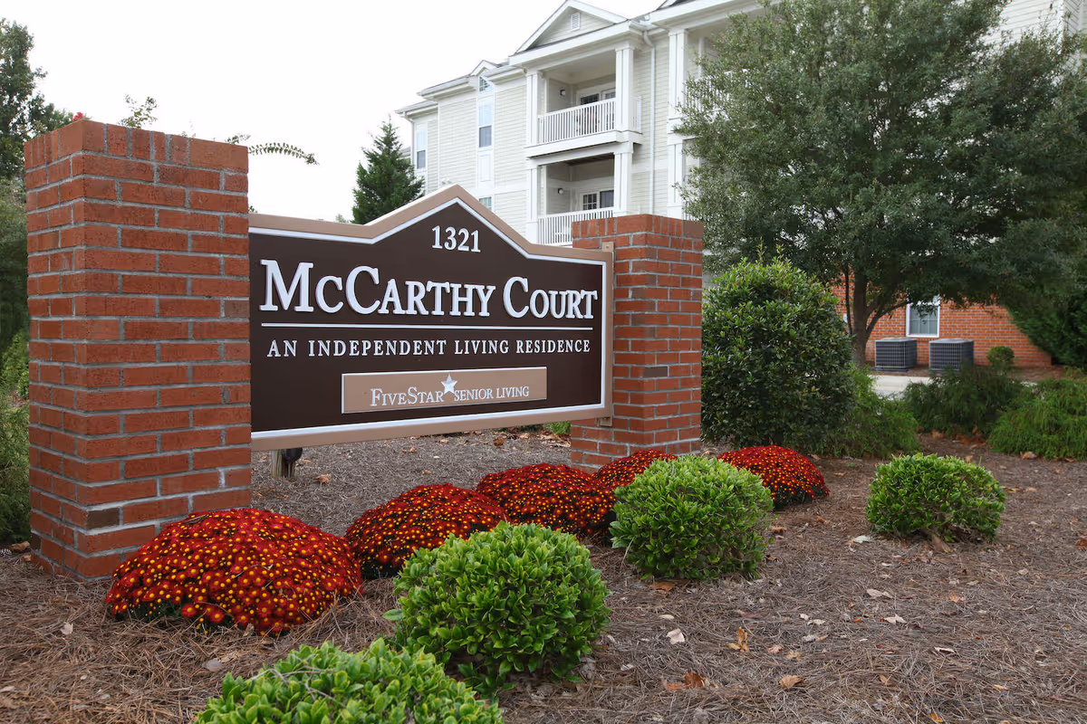A brick sign for McCarthy Court, an independent living residence, surrounded by neatly trimmed bushes and red flowers, with a multi-story residential building and trees in the background.