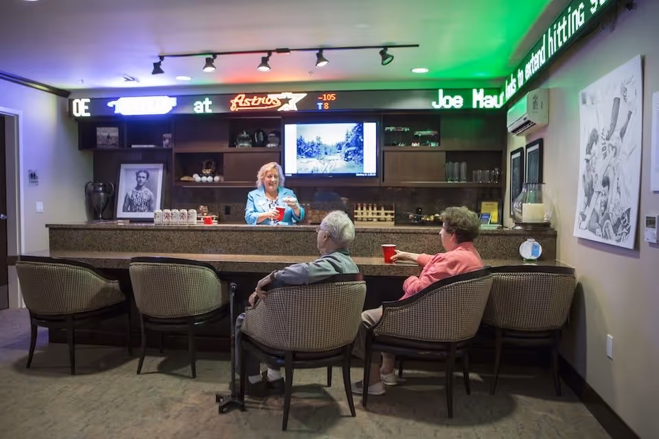 A cozy bar area inside a senior living facility with a woman standing behind the bar serving drinks to two elderly people seated on chairs. The bar has a granite countertop, shelves with decorative items, and a TV mounted on the wall. There is a neon sign with sports-related text above the bar and framed artwork on the walls.