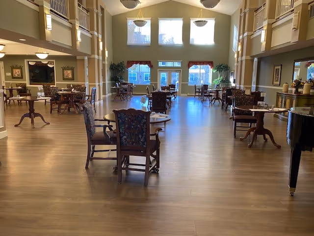 Spacious sunlit dining room with wooden floors, multiple dining tables and chairs, a piano, and high ceilings with upper-level balconies and large windows.