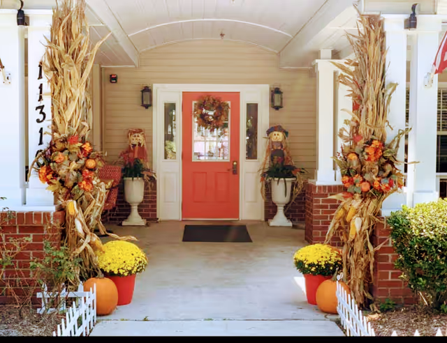 Front entrance with a red door decorated for fall, flanked by corn stalks, pumpkins, potted mums, and seasonal decorations.